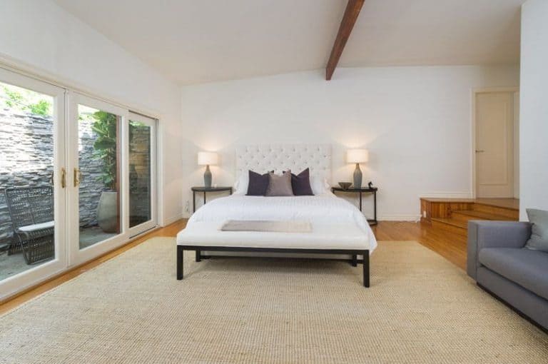 This master bedroom boasts a huge mat covering the hardwood flooring. It has a gray couch on the side. The bed is lighted by two table lights on the sides.