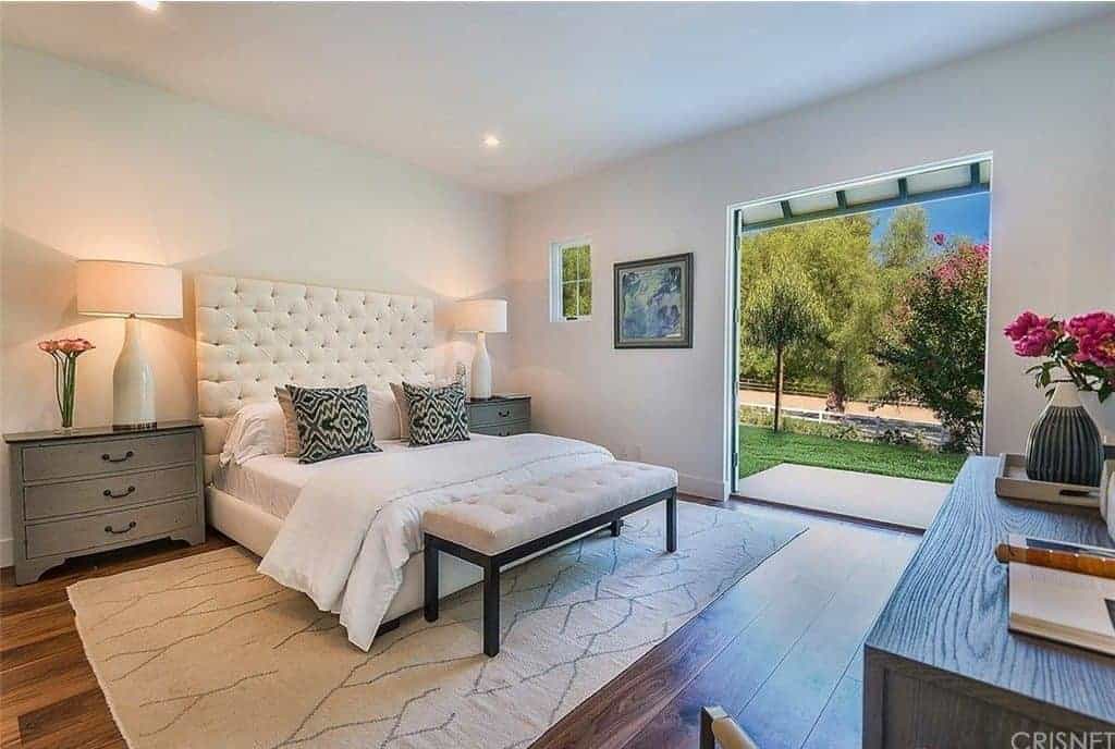 This master bedroom offers a high-class bed lighted by two table lamps on the sides. It has an area rug covering the room's hardwood flooring. There's an entryway leading the home terrace.