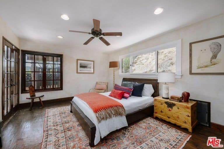 This master bedroom has a lot of wooden touch from the wooden-framed window to the hardwood flooring covered by a carpet with an Aztec design. It has a wooden chair, wooden side table, and a wooden bed frame.