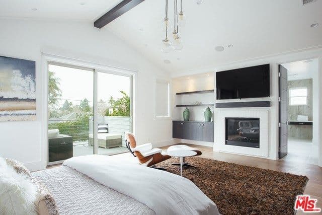 This master bedroom offers a large bed, a sitting area near the windows, a fireplace and a widescreen TV on top of it, lighted by wall sconces.