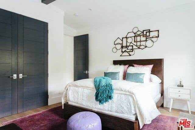 A master bedroom that has hardwood flooring covered by a carpet with a little purple chair. This room features a geometrical fixture hanging by the wall.