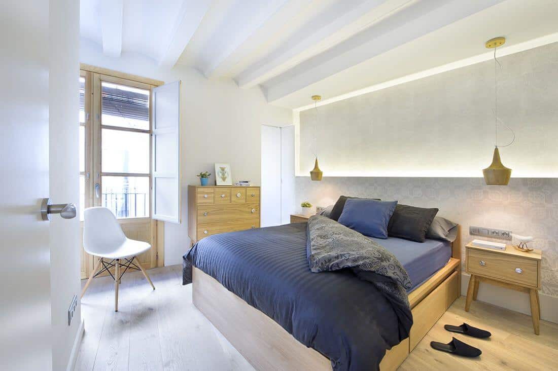 This master bedroom features hardwood flooring and a white classy wall. The bed highlights two side tables and lit by several pendant lights.