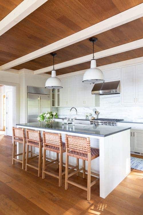 For a shabby chic style, this kitchen has a white island with a grey countertop. The woven four-legs chairs match the wooden flooring just right, while the white splashback and pantry match the inverted bowl hanging lights.