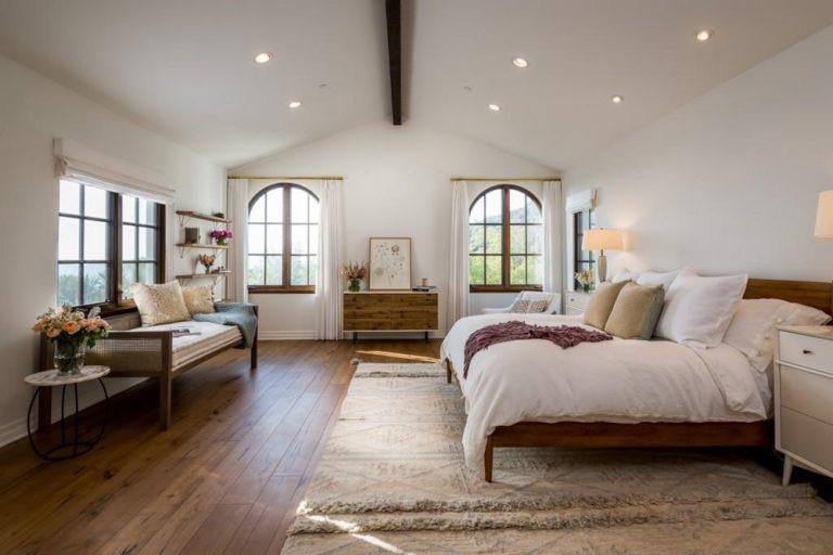 A comfortable master bedroom featuring hardwood flooring cover by a large carpet with white walls and ceiling.