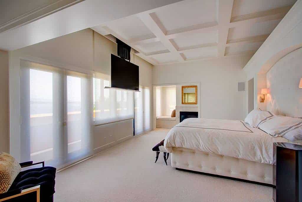 This white master bedroom features a large widescreen TV hanging from the ceiling in front of the large bed set lighted by two built-in lamps beside. The room offers a modern chair on the side.