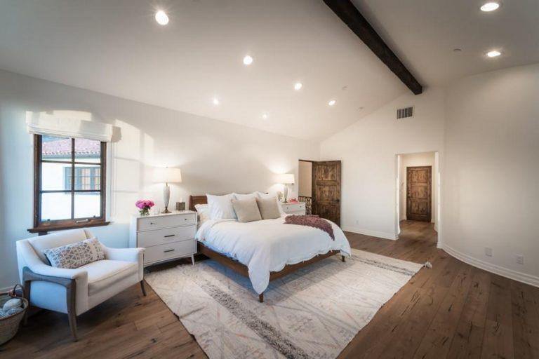 A spacious master bedroom featuring hardwood flooring topped by a large area rug along with white walls and a vaulted ceiling with an exposed beam.