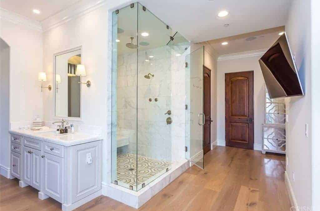 This primary bathroom contrasted with a white wood sink vanity that sits beside the walk-in shower enclosed in glass. It has a TV hanging on the wall in front of the walk-in shower.