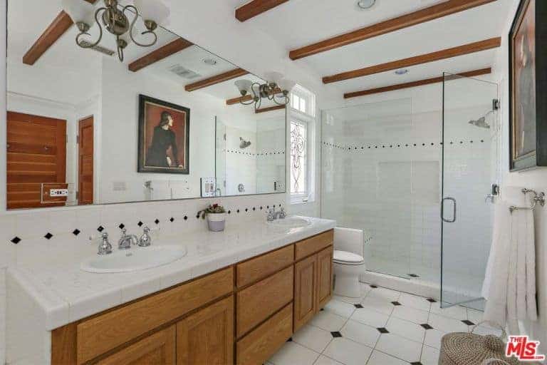 An enchanting portrait is reflecting in the frameless mirror mounted with vintage sconces in this primary bathroom with wood beam ceiling and white tiled flooring accented by the black diamond pattern.