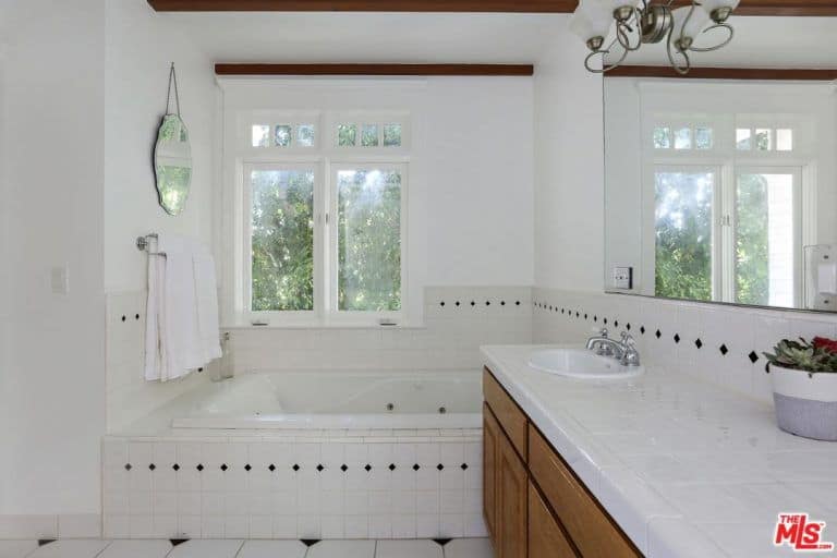 Small primary bathroom highlighting a drop in the bathtub next to the wooden vanity and underneath a white framed window bringing natural light in.