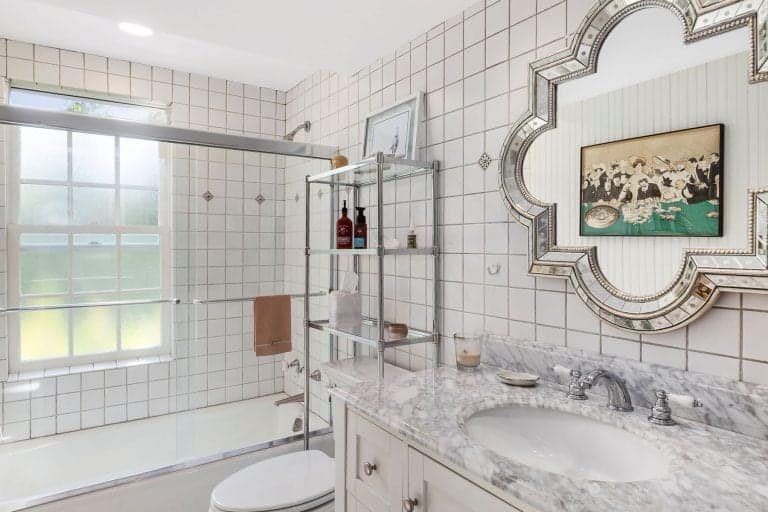 The small white bathroom has a marble countertop and a traditional toilet. It also has a tile wall mounted with wooden sink vanity.