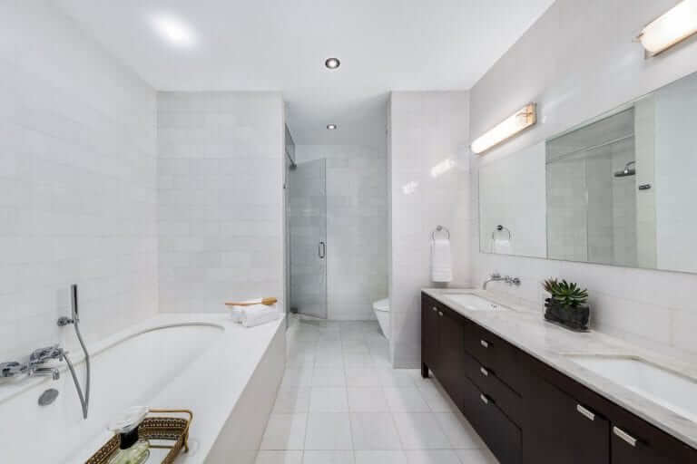 Darkwood vanity with a marble countertop and dual basin sinks is the highlights of this primary bathroom boasting a deep soaking tub and a large mirror pairing the white walls.