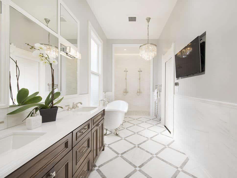 Modern bathroom featuring a freestanding tub and wooden cabinetry on the marble countertops with dual sink lightens by a stunning chandelier. This room also has TV on the white walls.