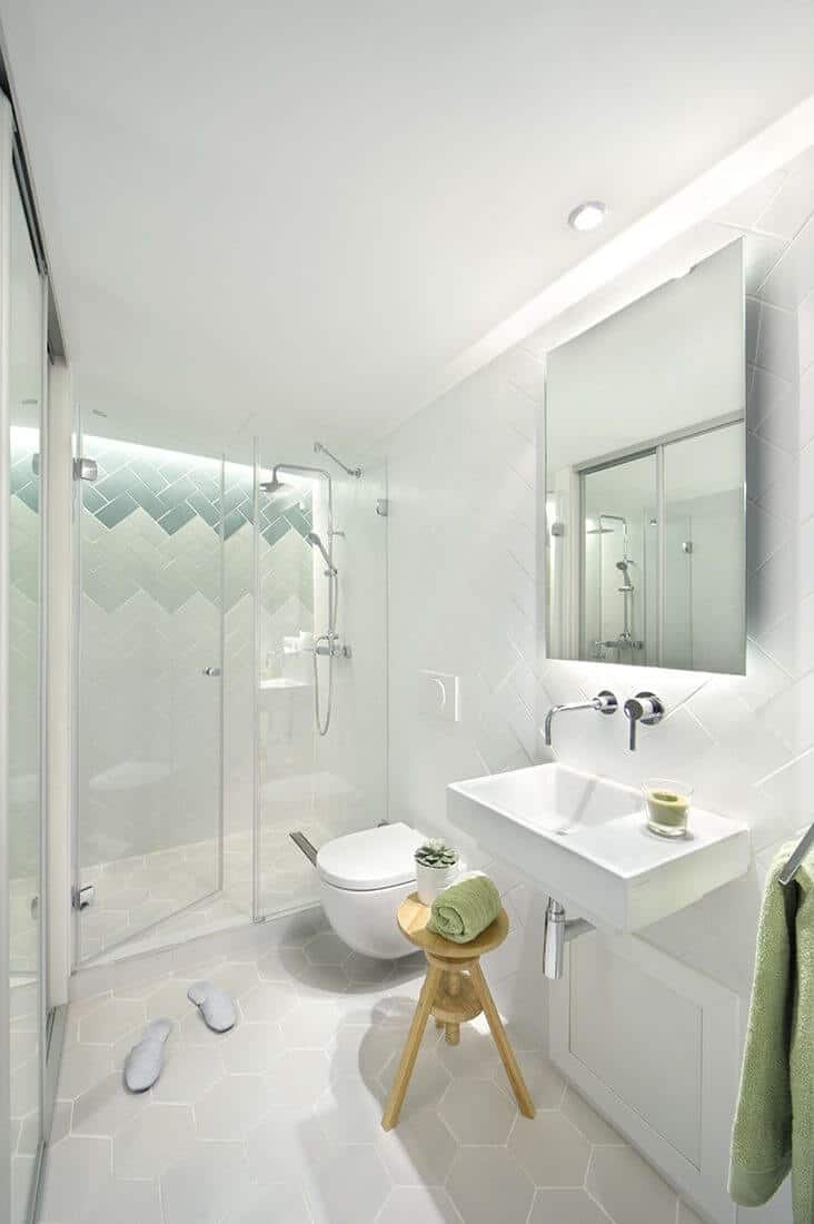White primary bathroom with a shower area and a small mirror over the basin sink. Modern bathroom with a wall-hung toilet and wooden chair beside.