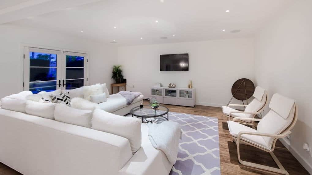 This charming white living room with an L-shape sofa set and glass table on top of a purple carpet covering the rustic hardwood flooring.