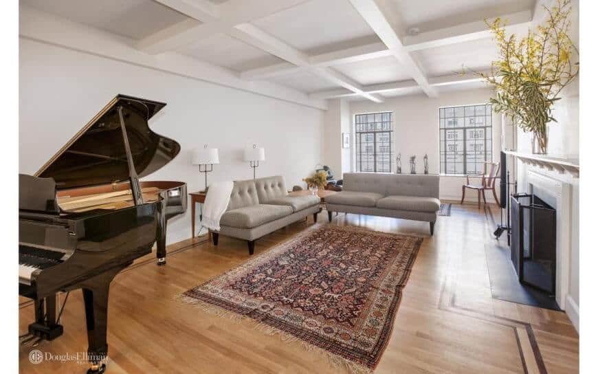 Spacious white formal living room featuring an elegant black piano, a cozy sofa set, and a fireplace. This room has hardwood flooring and elegant rug.