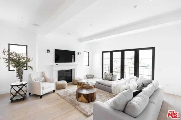 A white formal living room with cozy sofa set and a small table on the rug. This room has a fireplace and a TV on the wall.
