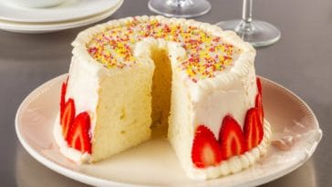 A sliced angel food cake with white frosting and garnish of strawberries and rainbow sprinkles.