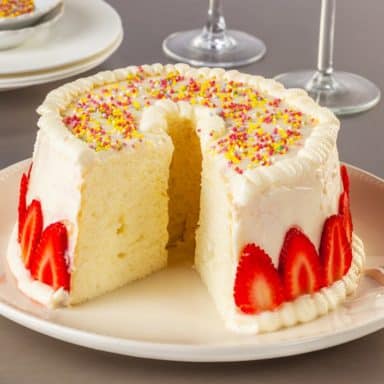 A sliced angel food cake with white frosting and garnish of strawberries and rainbow sprinkles.