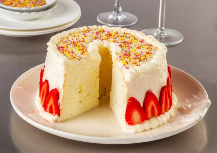A sliced angel food cake with white frosting and garnish of strawberries and rainbow sprinkles.