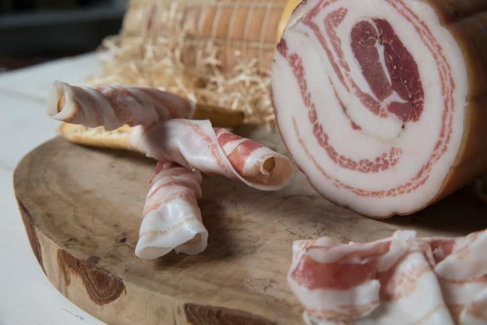 A close look at a slab of pancetta with slices on a chopping board.