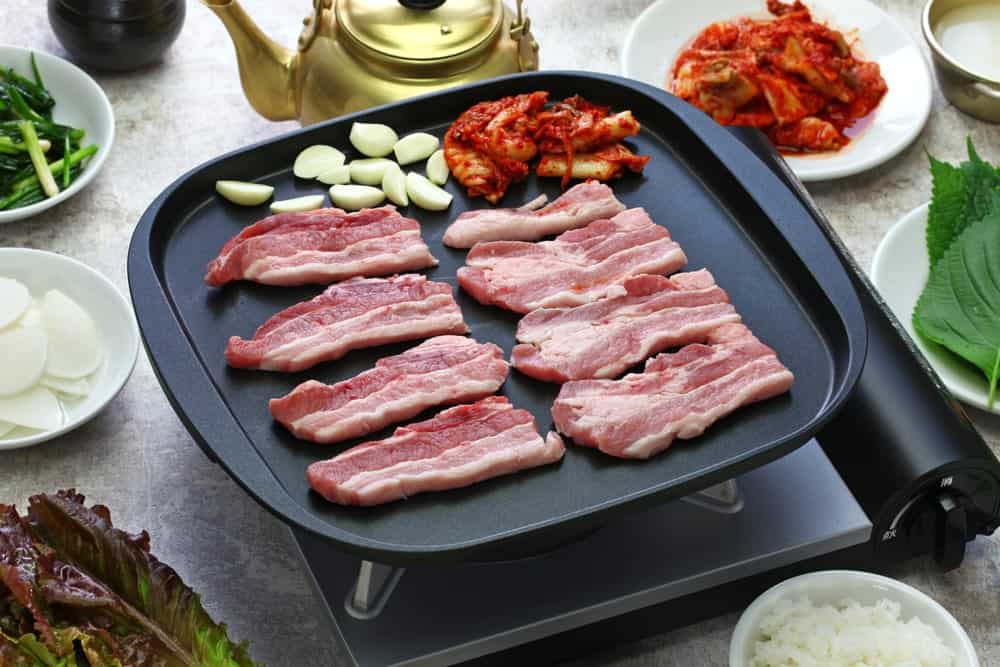 A close look at Korean Samgyeopsal bacon strips on a frying pan with side dishes.