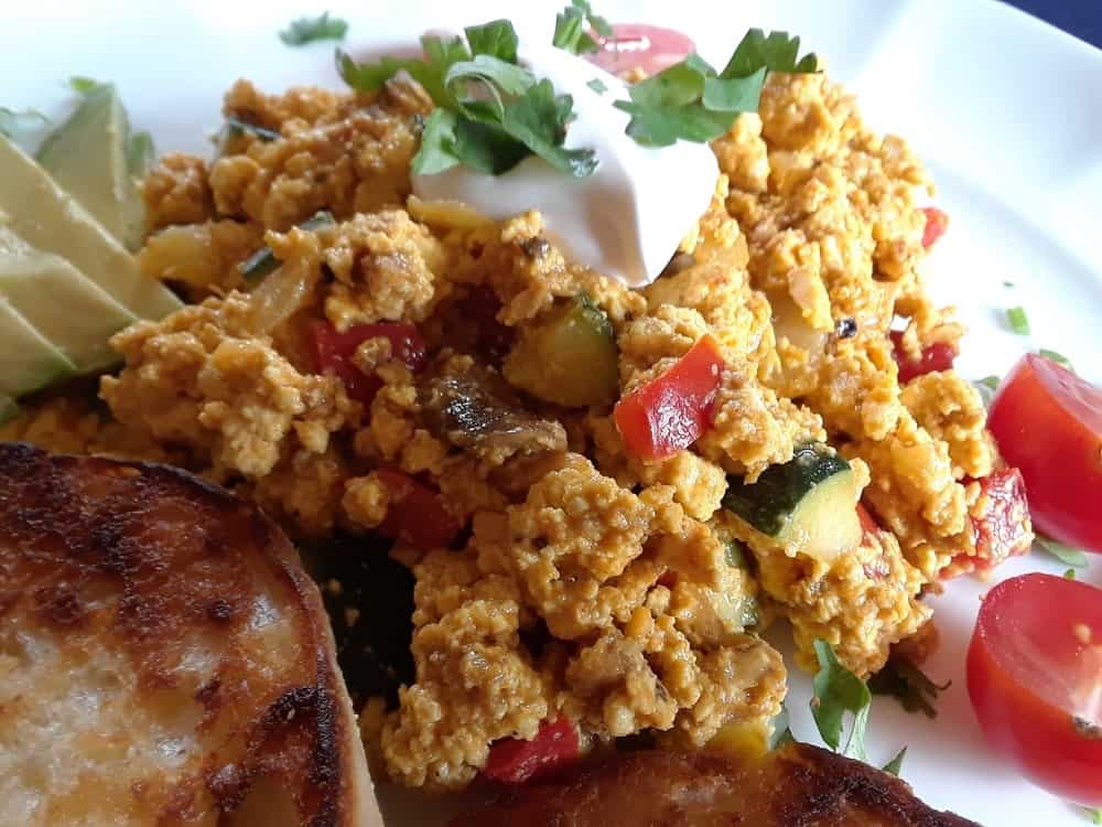 A plate of freshly-cooked vegan tofu scramble with a side of bread, avocado and topped with a cheese dressing.