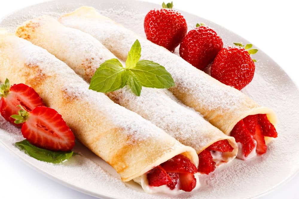 A plate of strawberry cream cheese crepes with fresh strawberry on the side.