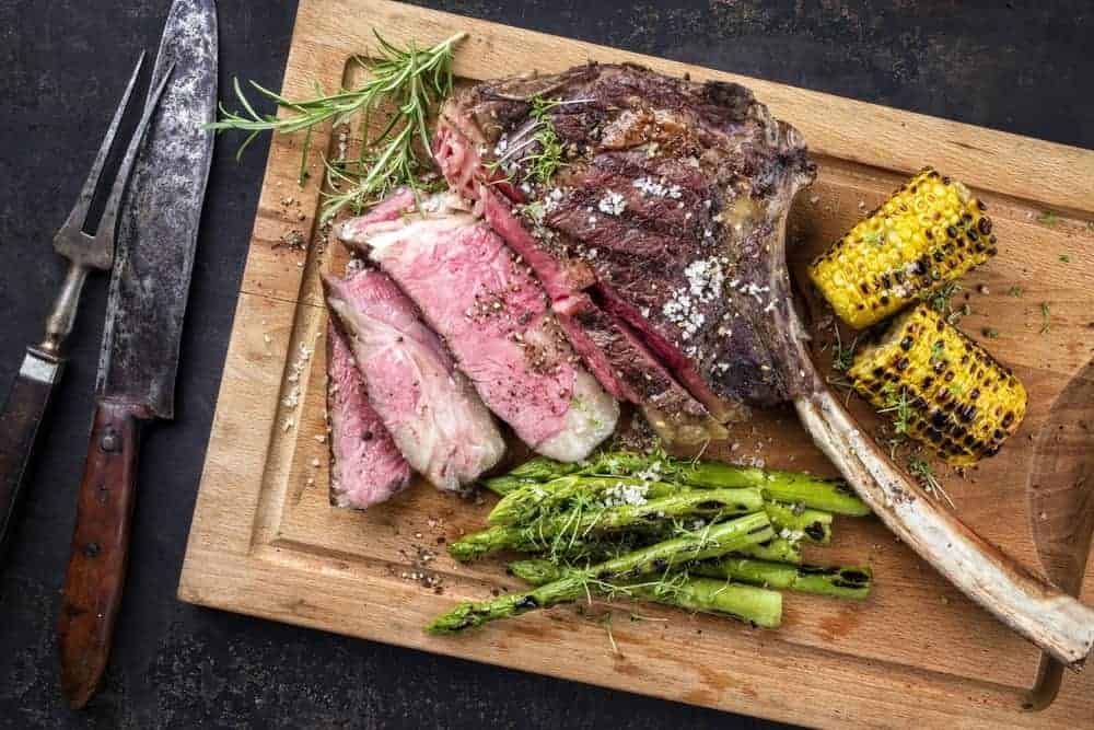 Sliced wagyu tomahawk steak with corns and asparagus on wooden chopping board.
